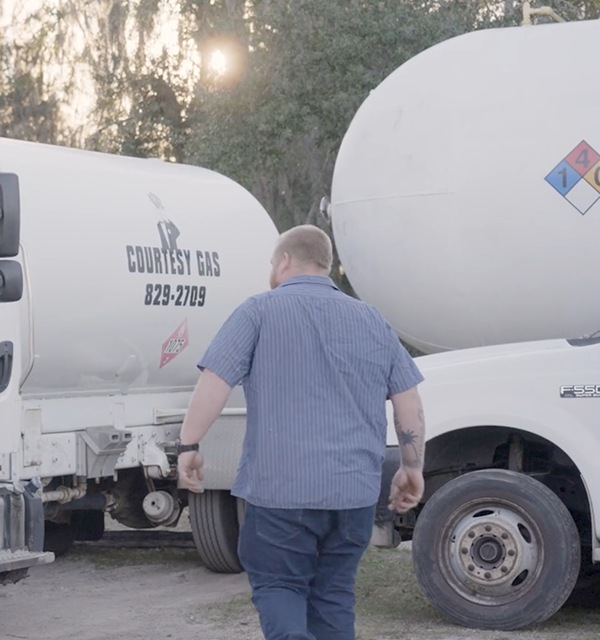 Consulting firm real client of Cogent Analytics- Male Professional in Blue Striped Shirt Walking Near Gas Trucks in Industrial Setting with Trees and Sunlight