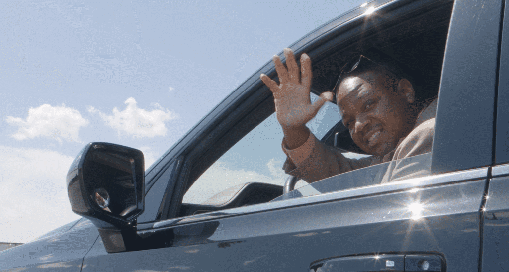 A black male driver dressed in a suit waving out the window of a limo.