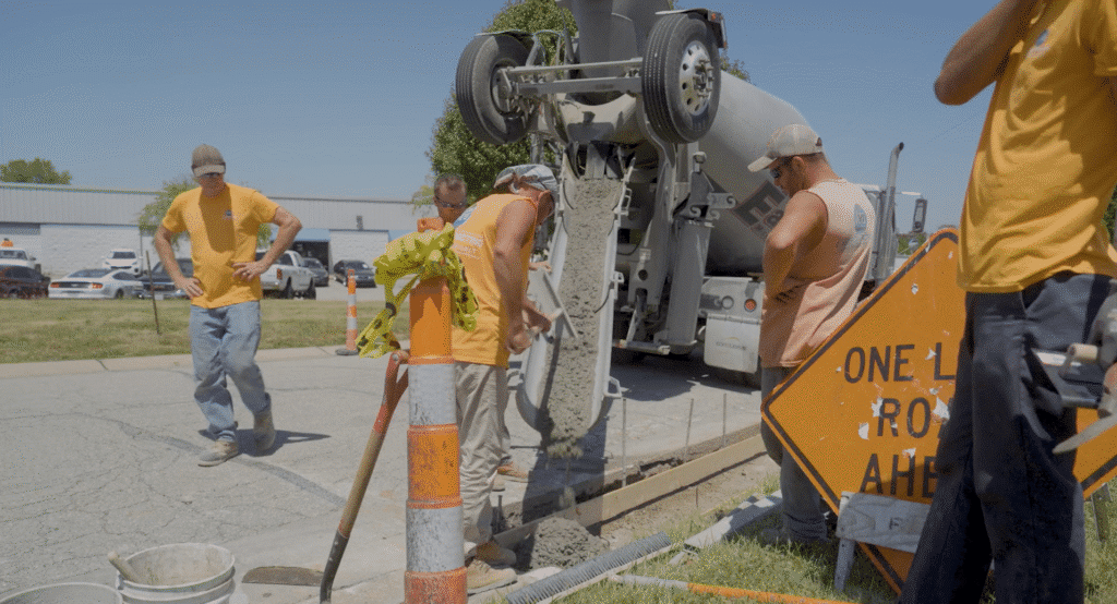Workers in orange shirts pour and level wet concrete from a cement truck into a roadside trench.
