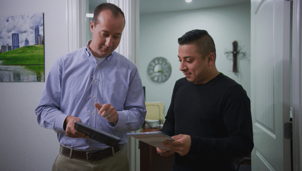 Two colleagues in a bright hallway reviewing information on a digital tablet and a printed document.