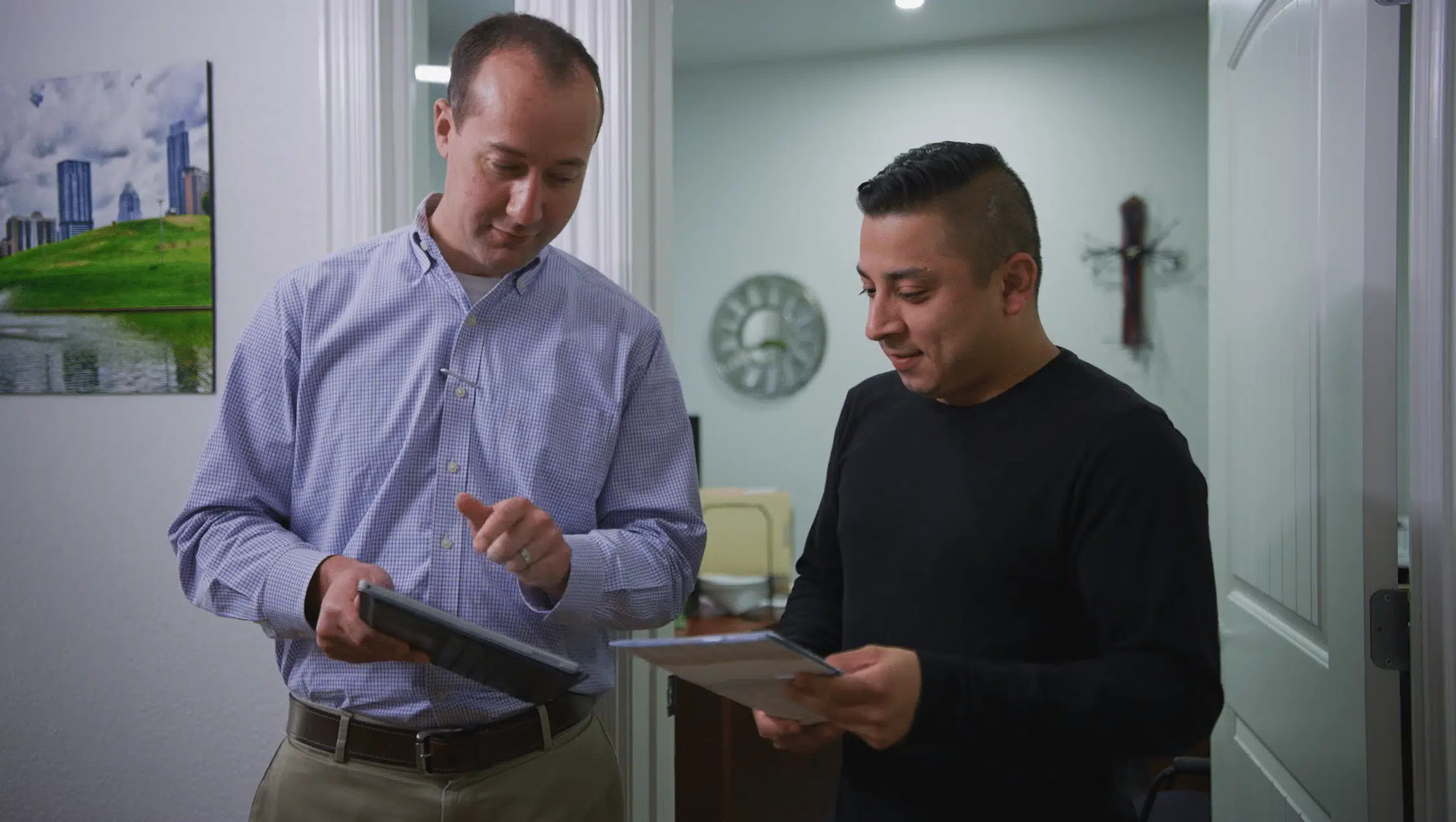 Two colleagues in a bright hallway reviewing information on a digital tablet and a printed document.
