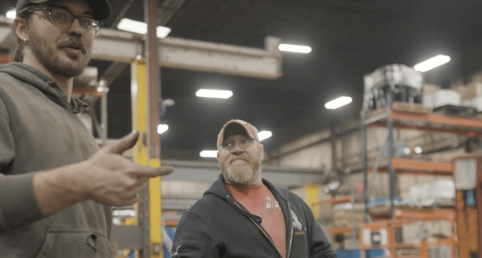 A young man in a grey hoodie and glasses gestures while speaking to a bearded man in a red shirt and black zip-up hoodie in a large industrial warehouse.