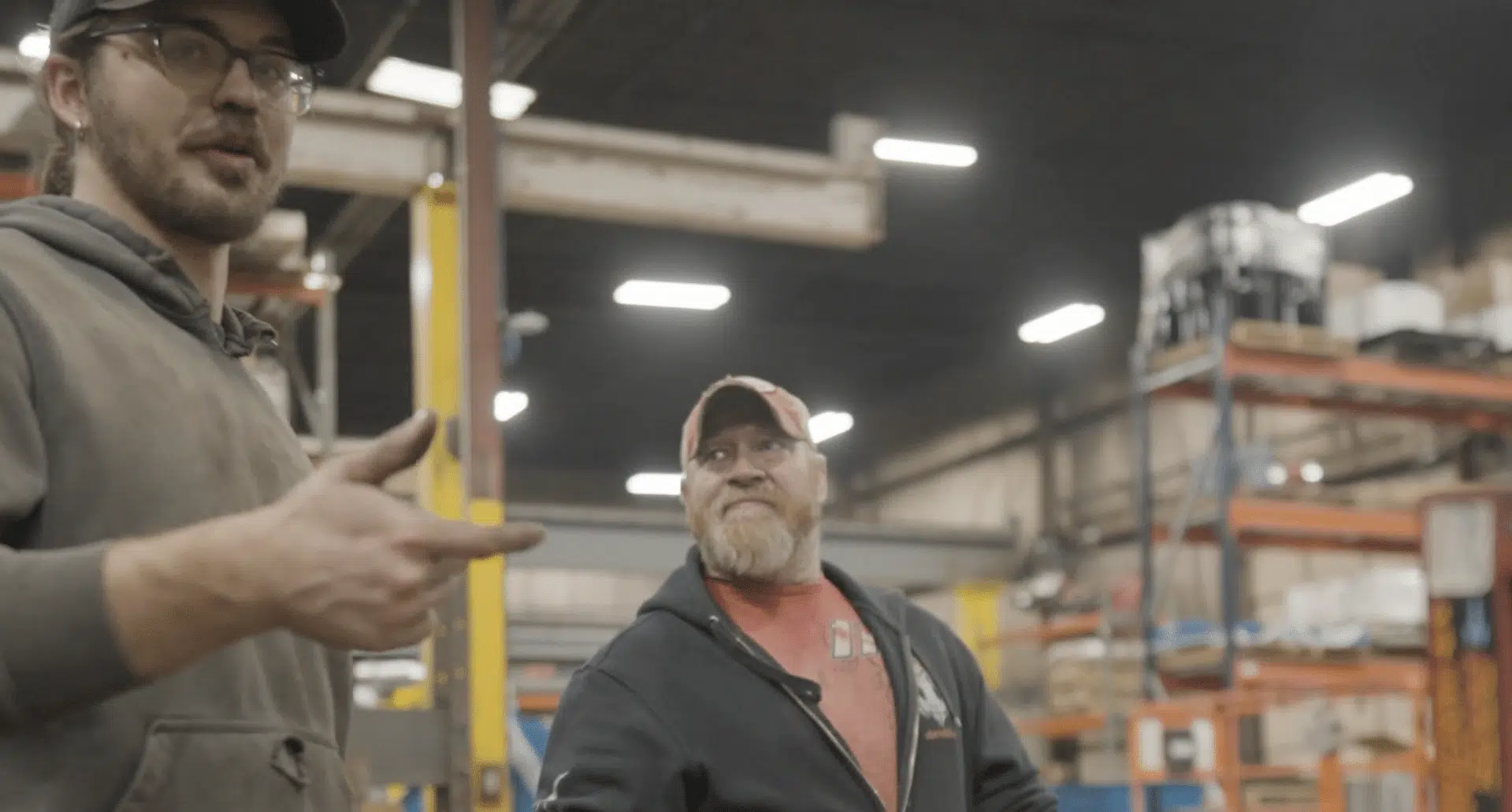 A young man in a grey hoodie and glasses gestures while speaking to a bearded man in a red shirt and black zip-up hoodie in a large industrial warehouse.