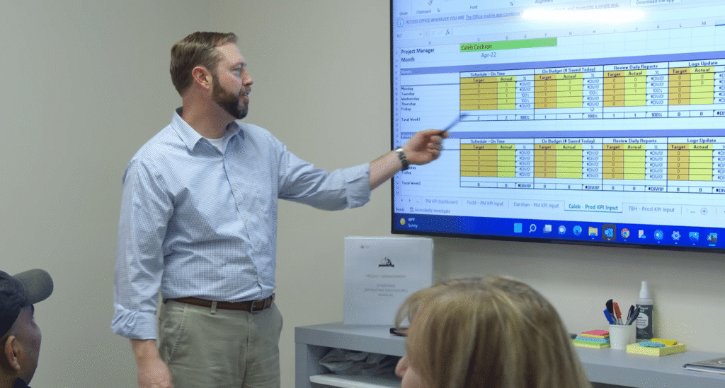 A man pointing a pen toward a project management spreadsheet displayed on a large wall monitor during a meeting.