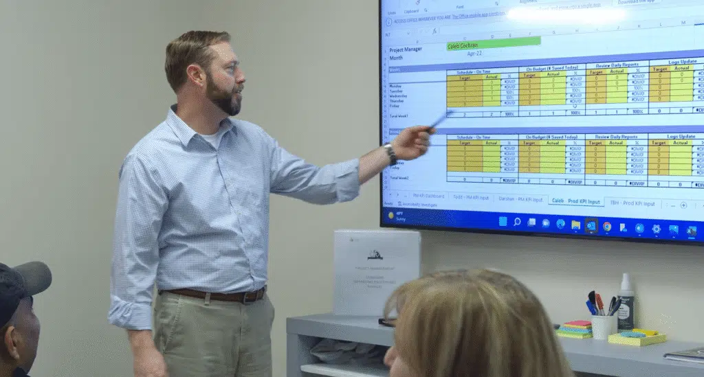 A man pointing a pen toward a project management spreadsheet displayed on a large wall monitor during a meeting.