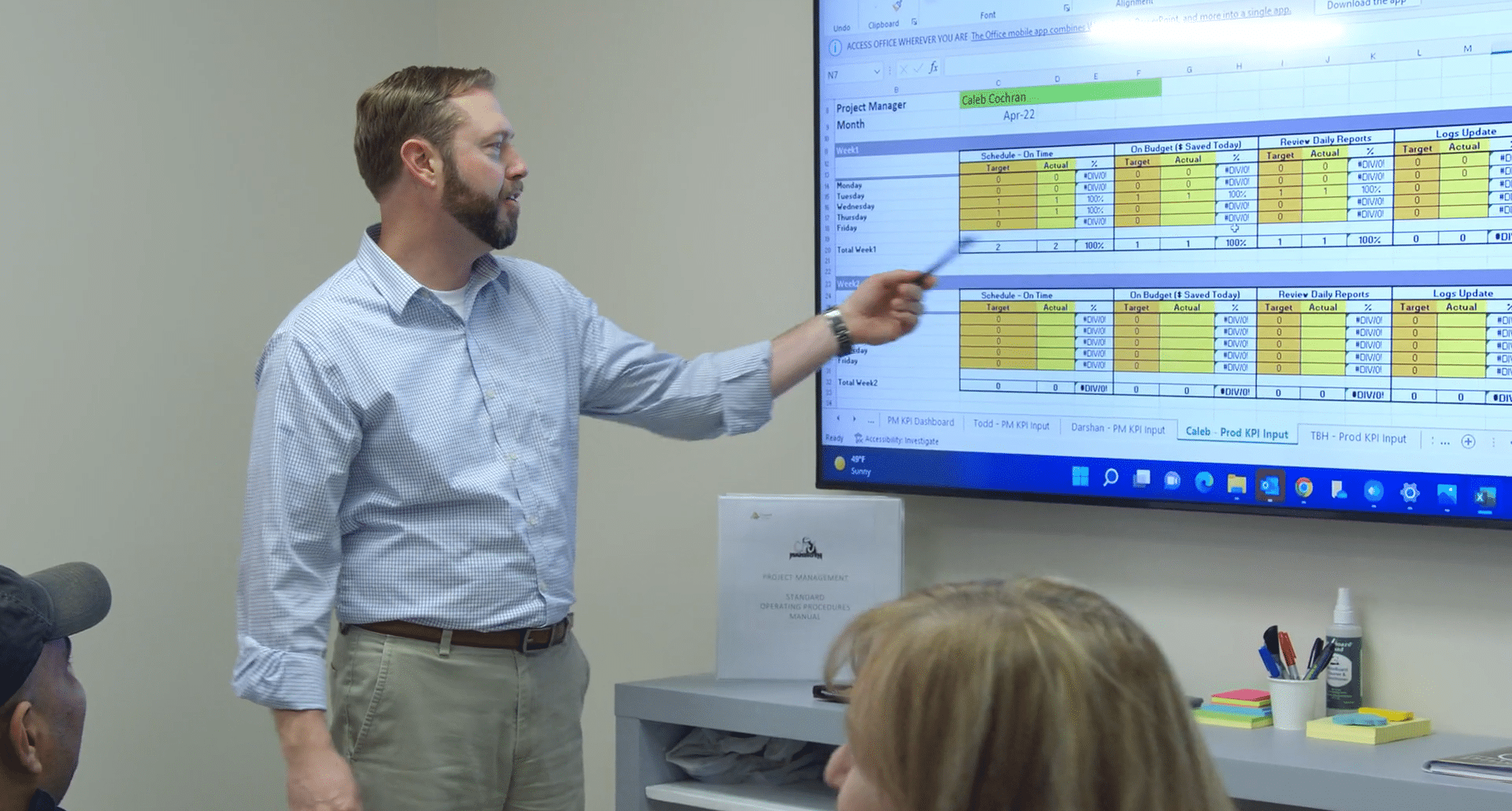 A man pointing a pen toward a project management spreadsheet displayed on a large wall monitor during a meeting.