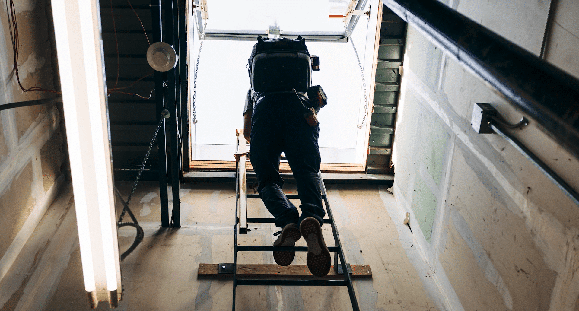 A low-angle view of a worker with a large equipment backpack climbing a metal ladder toward a bright roof hatch in an unfinished utility space.