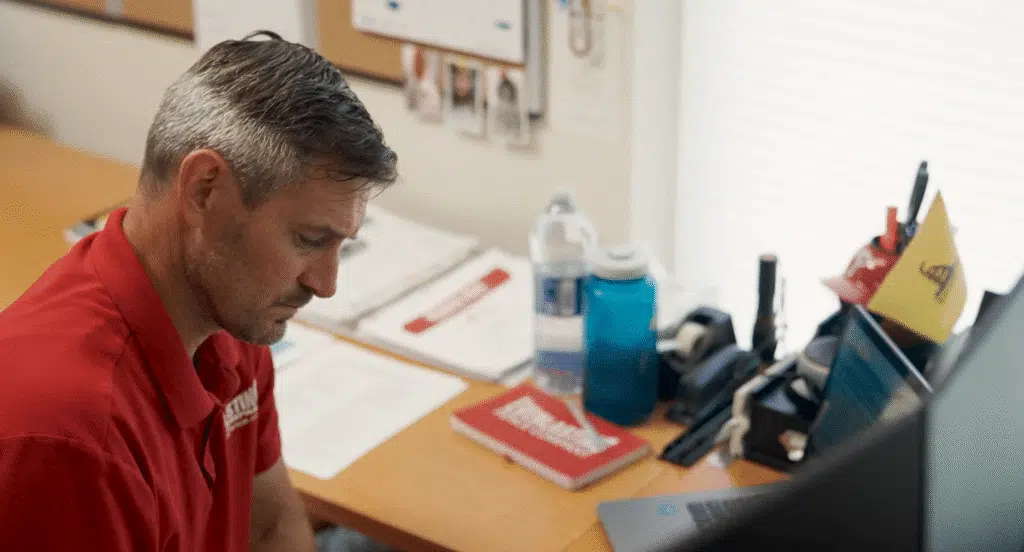 A man with short, graying hair wearing a red polo shirt sits at a wooden desk and focuses intently on paperwork and a laptop.