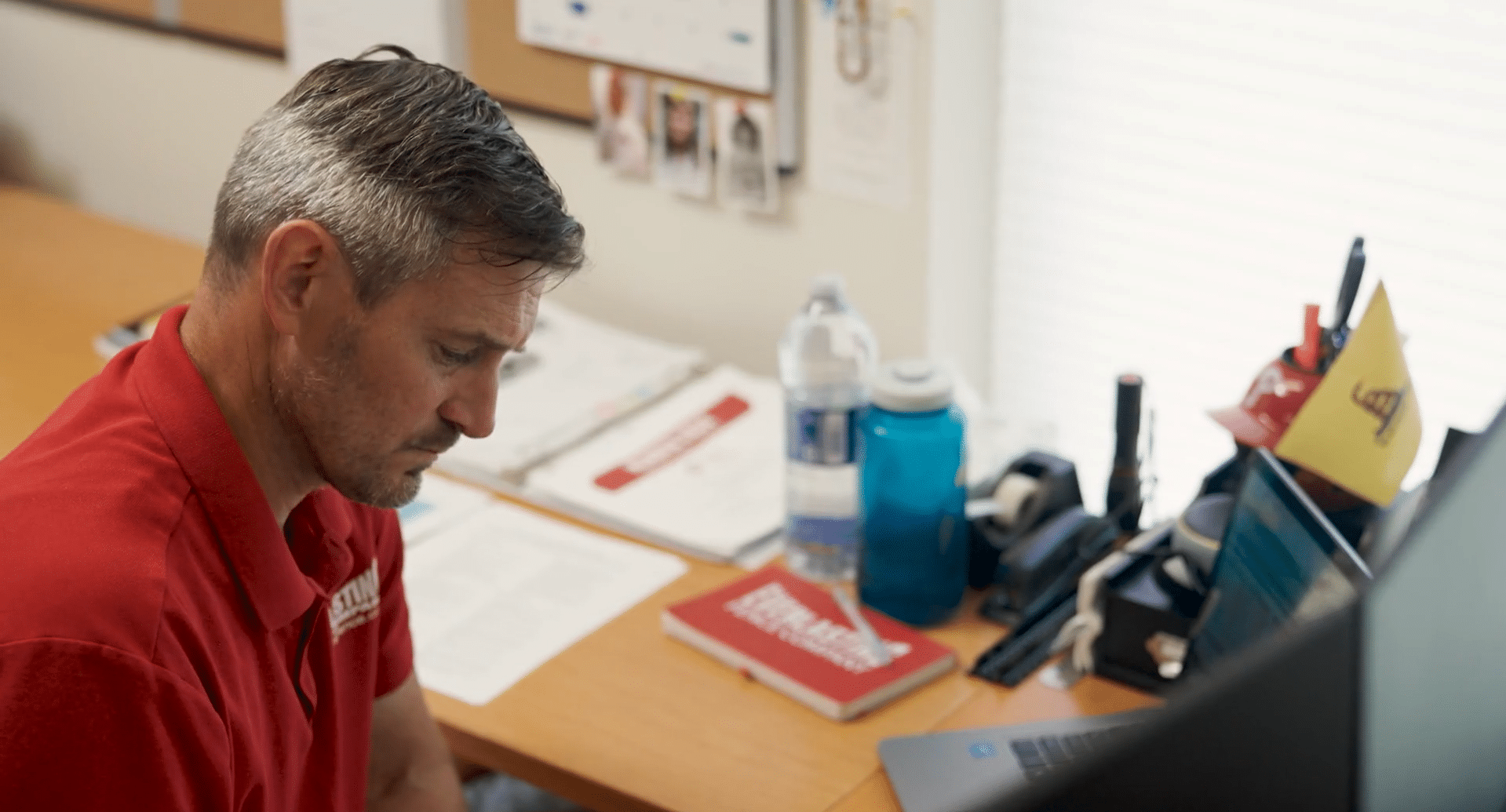 A man with short, graying hair wearing a red polo shirt sits at a wooden desk and focuses intently on paperwork and a laptop.