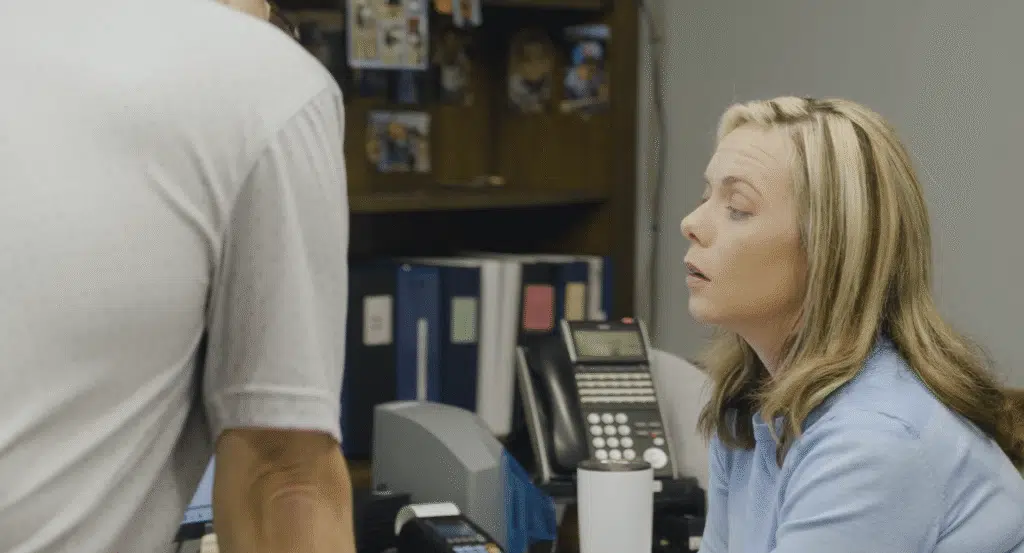 A woman in a blue sweater sits at an office desk and looks up to engage in a conversation with a colleague standing just out of frame.