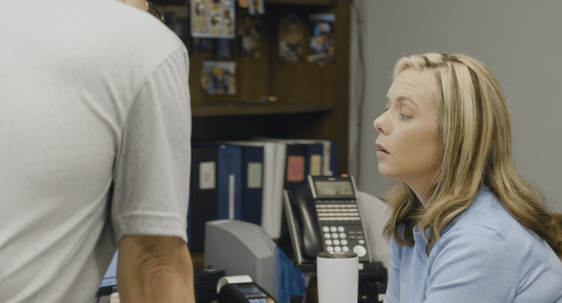 A woman in a blue sweater sits at an office desk and looks up to engage in a conversation with a colleague standing just out of frame.