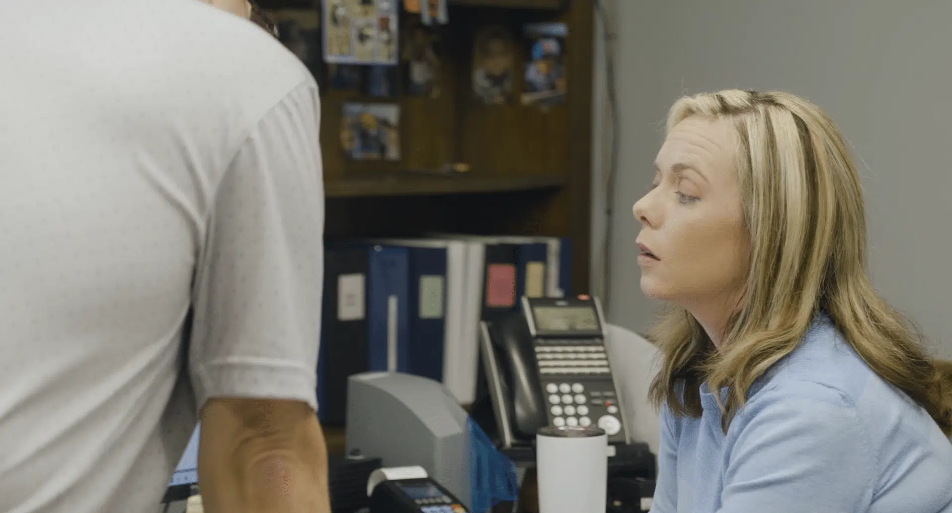 A woman in a blue sweater sits at an office desk and looks up to engage in a conversation with a colleague standing just out of frame.