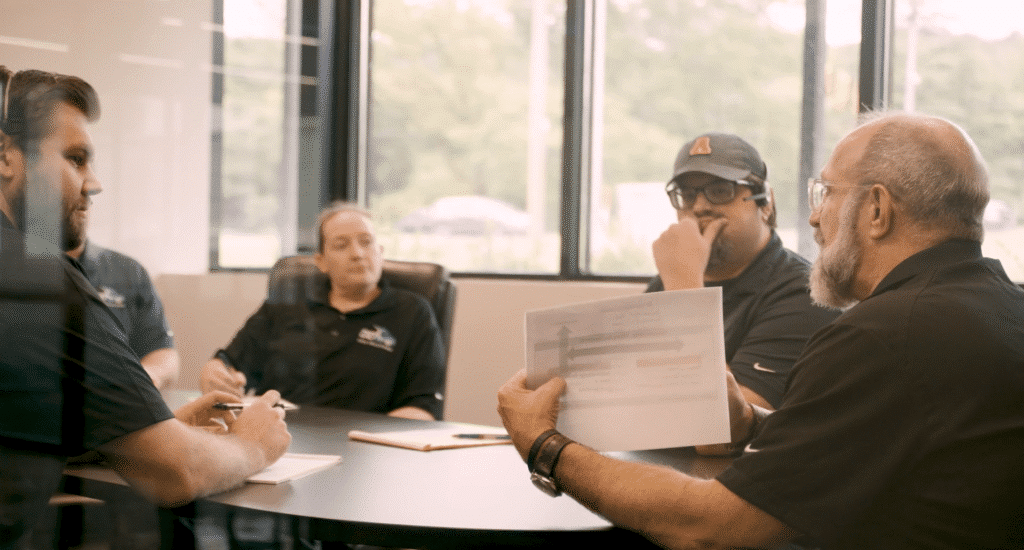 Four professionals gather around a table in a bright conference room for a meeting; one man holds up a document with a diagram to show the group.