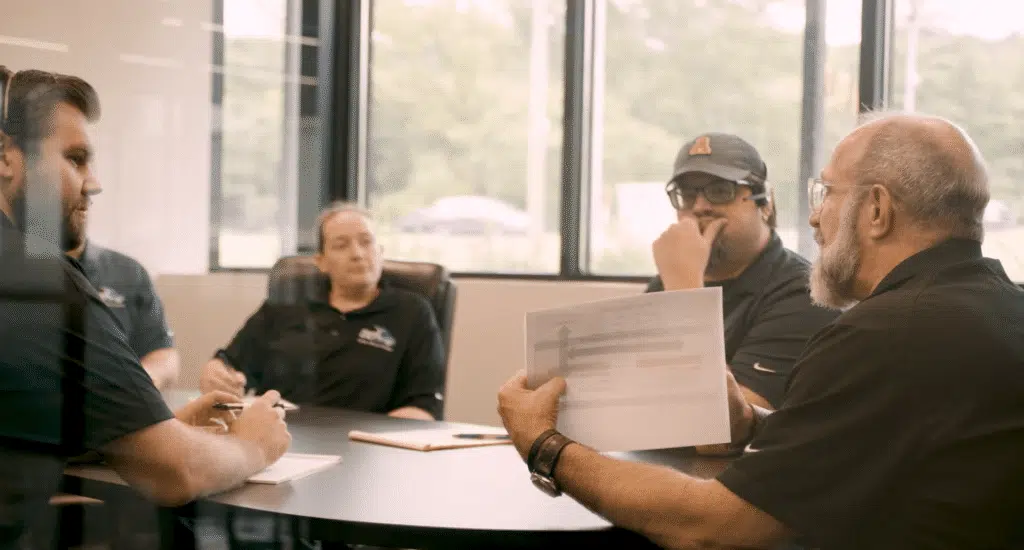 Four professionals gather around a table in a bright conference room for a meeting; one man holds up a document with a diagram to show the group.