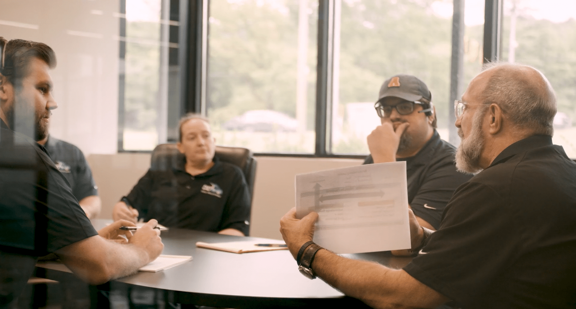 Four professionals gather around a table in a bright conference room for a meeting; one man holds up a document with a diagram to show the group.