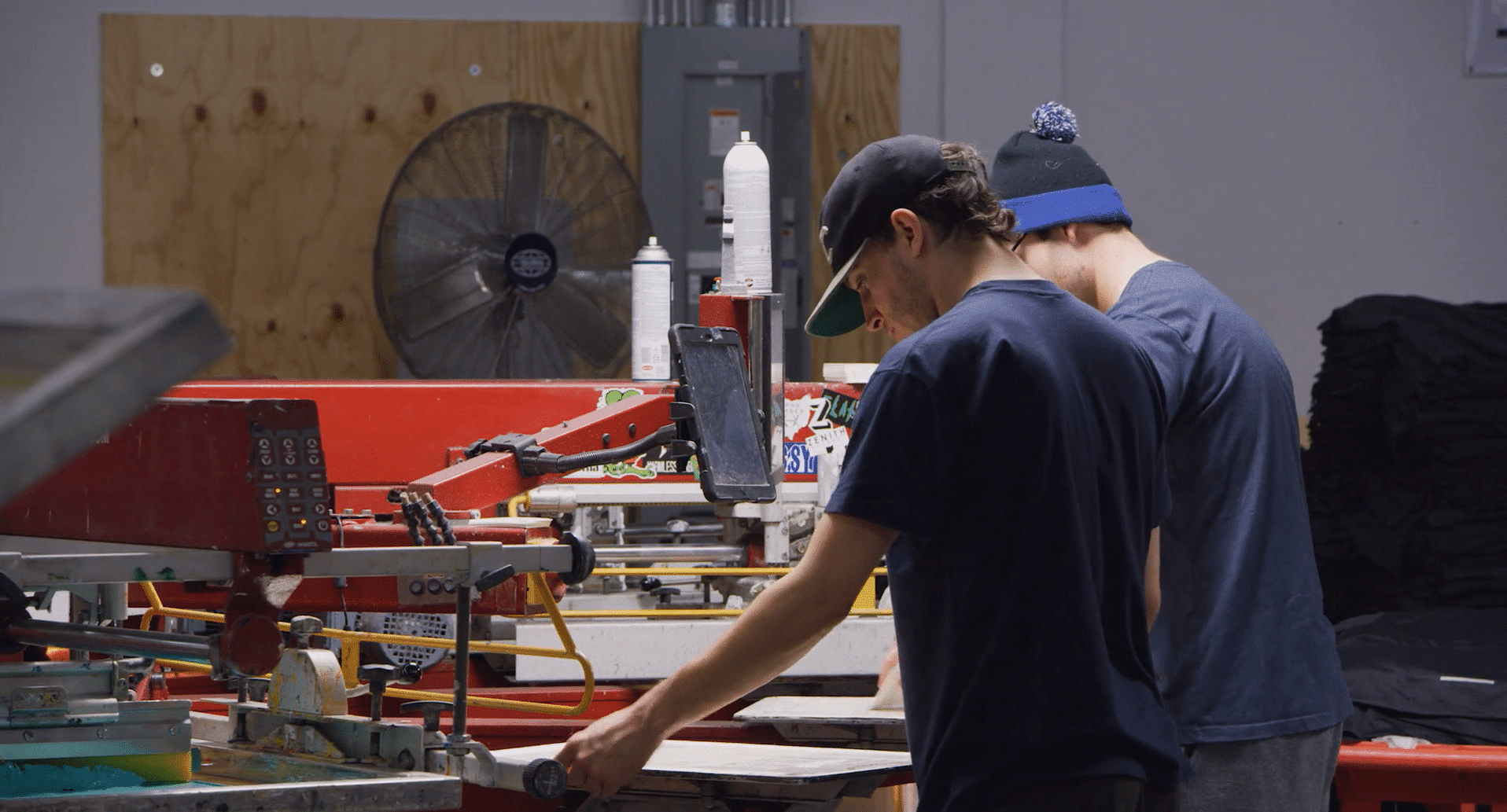 Two men in hats operate a red industrial screen-printing machine in a workshop setting.