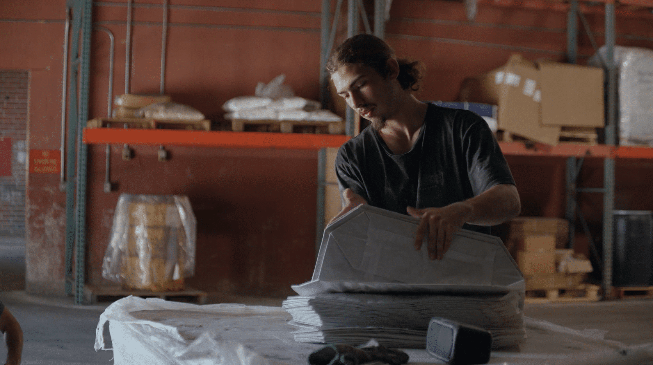 A young man with long hair tied back stands in a warehouse and carefully folds or organizes a stack of large white paper bags on a flat surface.
