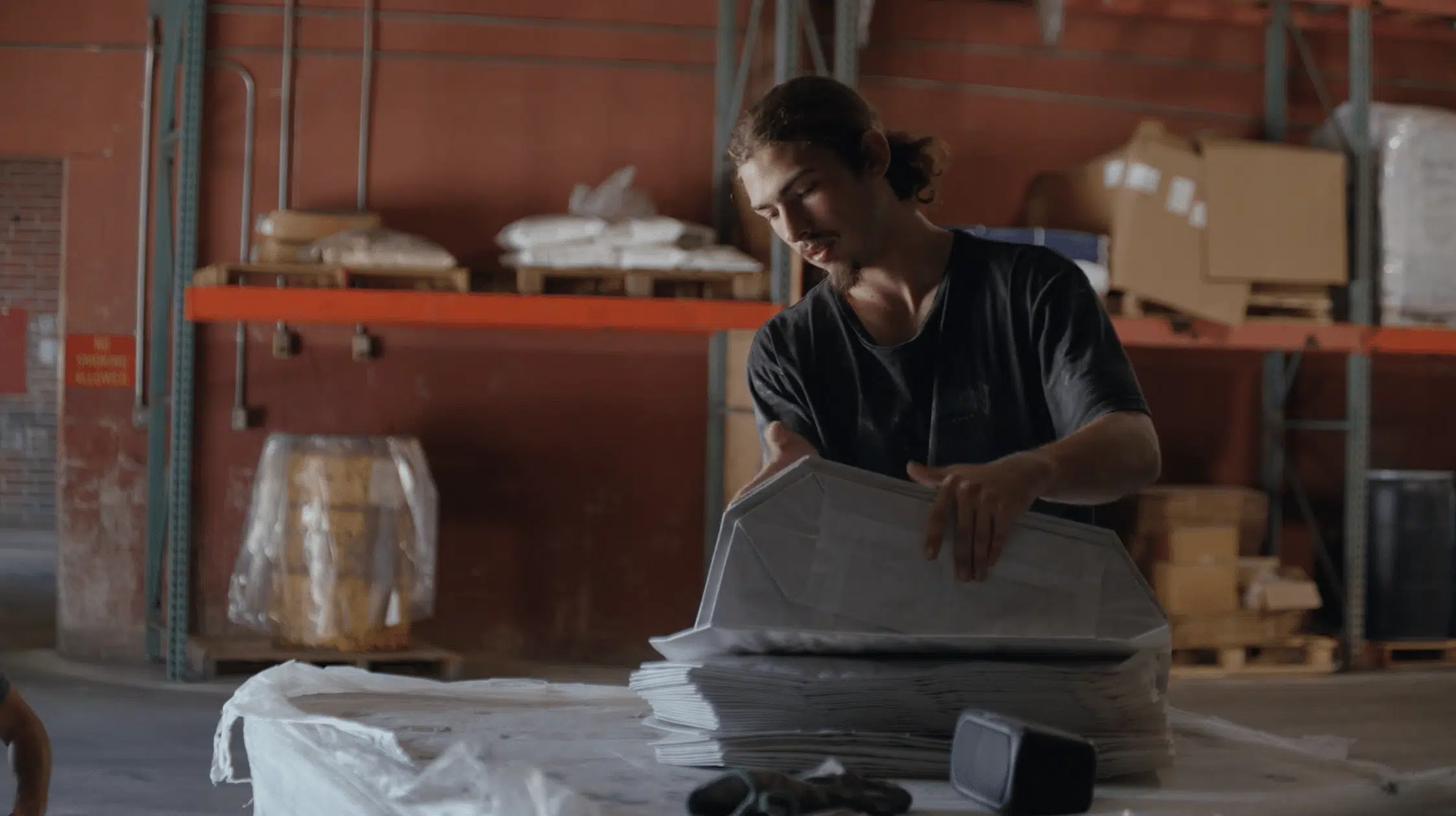 A young man with long hair tied back stands in a warehouse and carefully folds or organizes a stack of large white paper bags on a flat surface.
