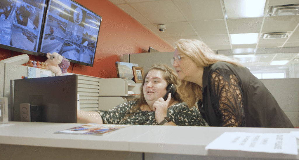 A woman in a floral print top speaks on a desk phone while a colleague in a black blouse leans in to look at a computer monitor displaying security camera feeds.