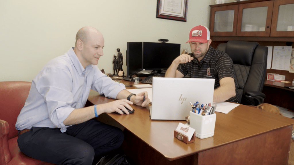 Two professionals seated at an office desk reviewing information on a laptop during a meeting, with office equipment and documents visible in the background.