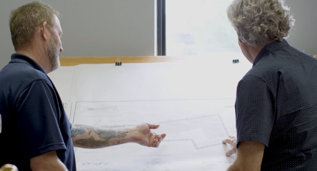 Two men, seen from behind, stand at a drafting table and point to a large architectural blueprint pinned to the board.