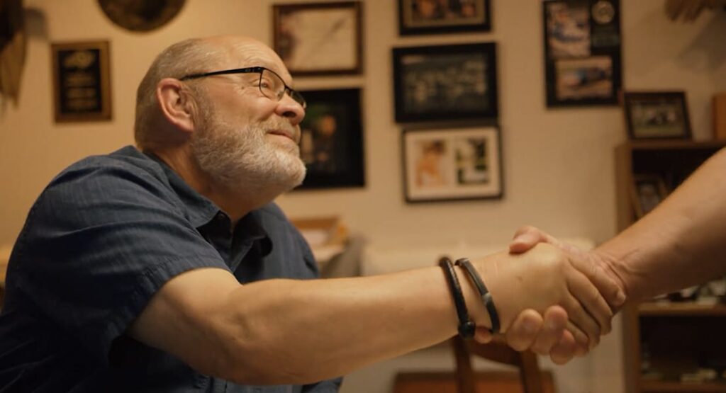 A smiling, older man with glasses and a gray beard shakes hands with another person in an office decorated with framed photos and plaques.
