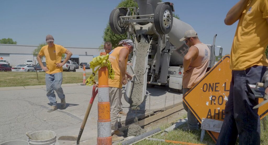 Construction workers in yellow shirts oversee a cement mixer truck pouring wet concrete into a roadside trench behind an orange warning sign.
