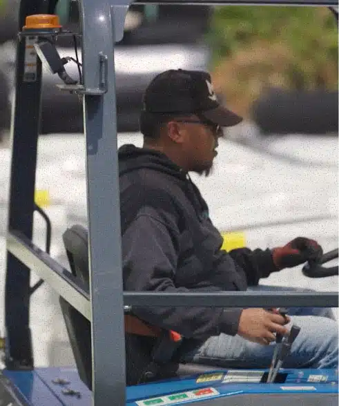 A man wearing a black baseball cap, sunglasses, and a dark hoodie sits in the driver's seat of a blue forklift.