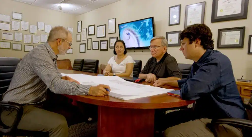 Four professionals sit around a large oval wooden table in a conference room, reviewing architectural blueprints together. Top Business Consulting Firms