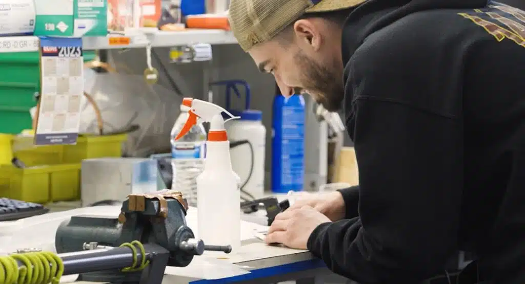 A man in a black hoodie and tan baseball cap leans over a workbench to focus on a precise task next to a white spray bottle and a metal vise.