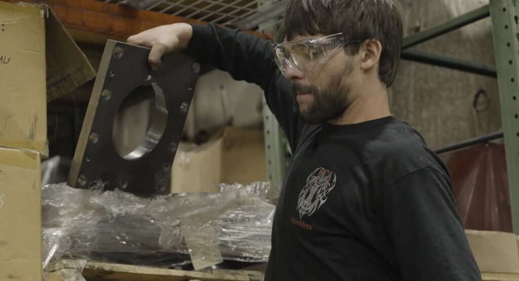 A man with a beard and safety glasses examines a heavy, square metal plate with a large circular hole in the center while standing in a warehouse.