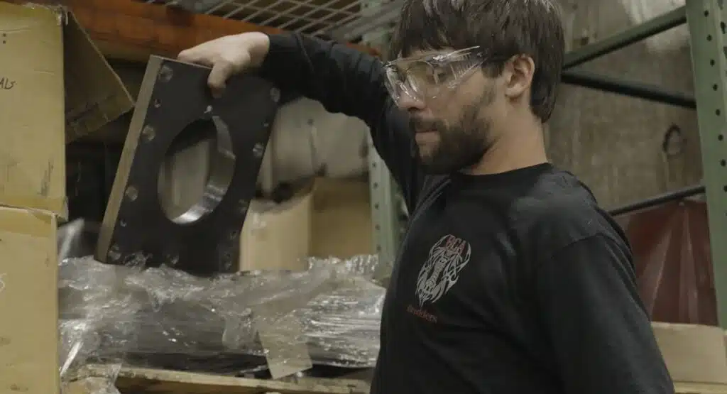 A man with a beard and safety glasses examines a heavy, square metal plate with a large circular hole in the center while standing in a warehouse.