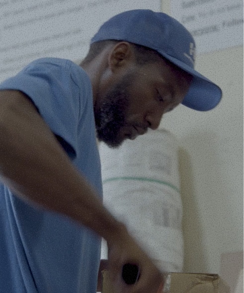 A man with a beard wearing a blue t-shirt and a blue baseball cap leans forward to focus on packing or opening a cardboard box.