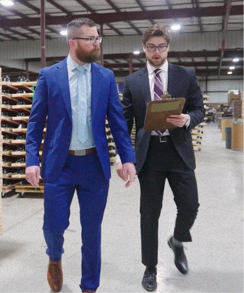 Two men in professional suits walk through a warehouse; one man in a dark suit reviews a clipboard while the other in a bright blue suit looks on.