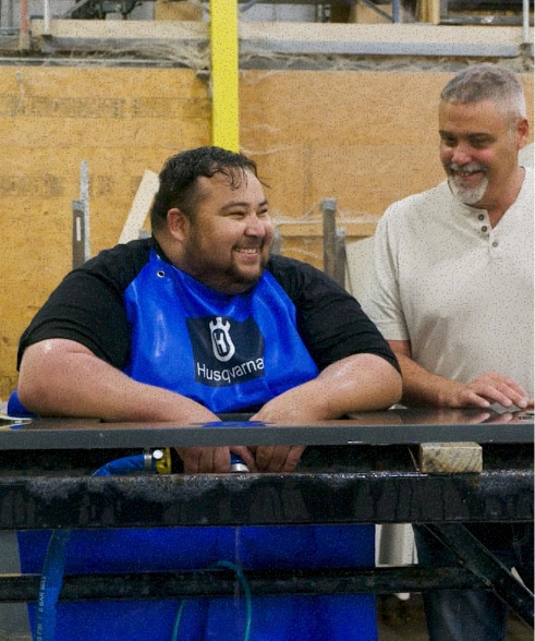 A smiling man in a blue Husqvarna apron leans against a stone countertop in a workshop while another man stands beside him.