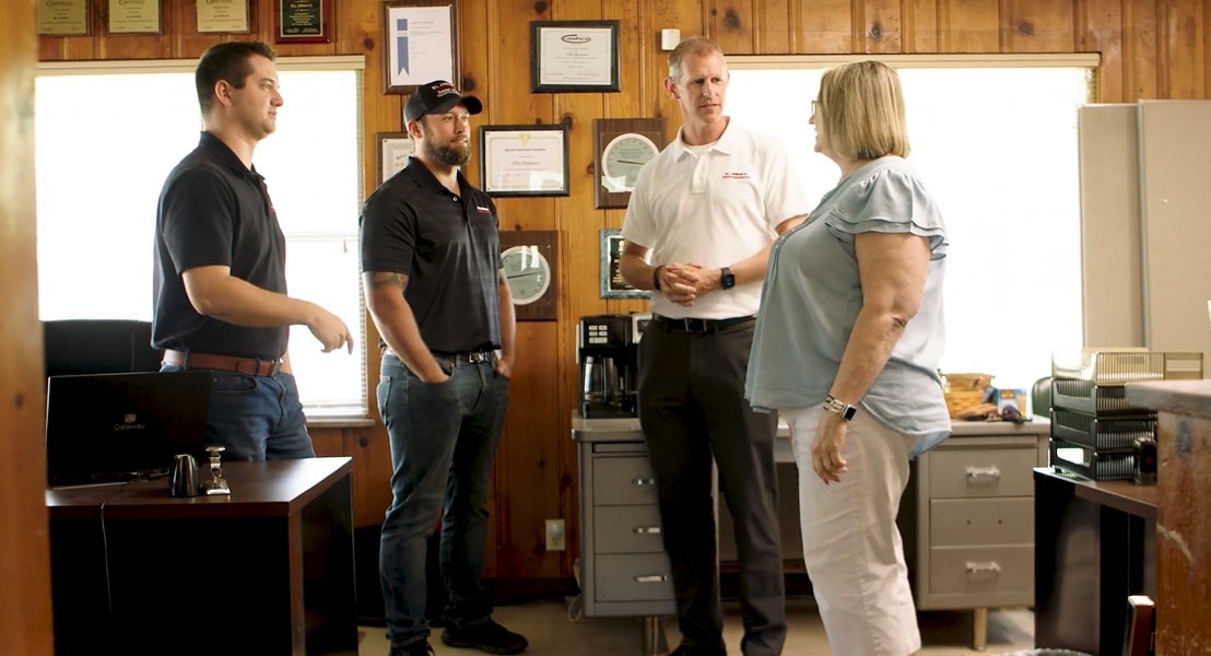 A woman in a light blue blouse speaks with three men in a wood-paneled office filled with framed certificates and professional awards. Business transition
