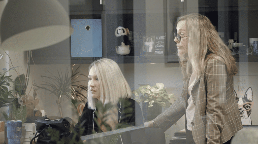 Two women work together in a modern office, seen through a glass wall with reflections. One woman sits at a desk looking at a computer while the other stands beside her, leaning in; shelves, plants, and personal items fill the background.