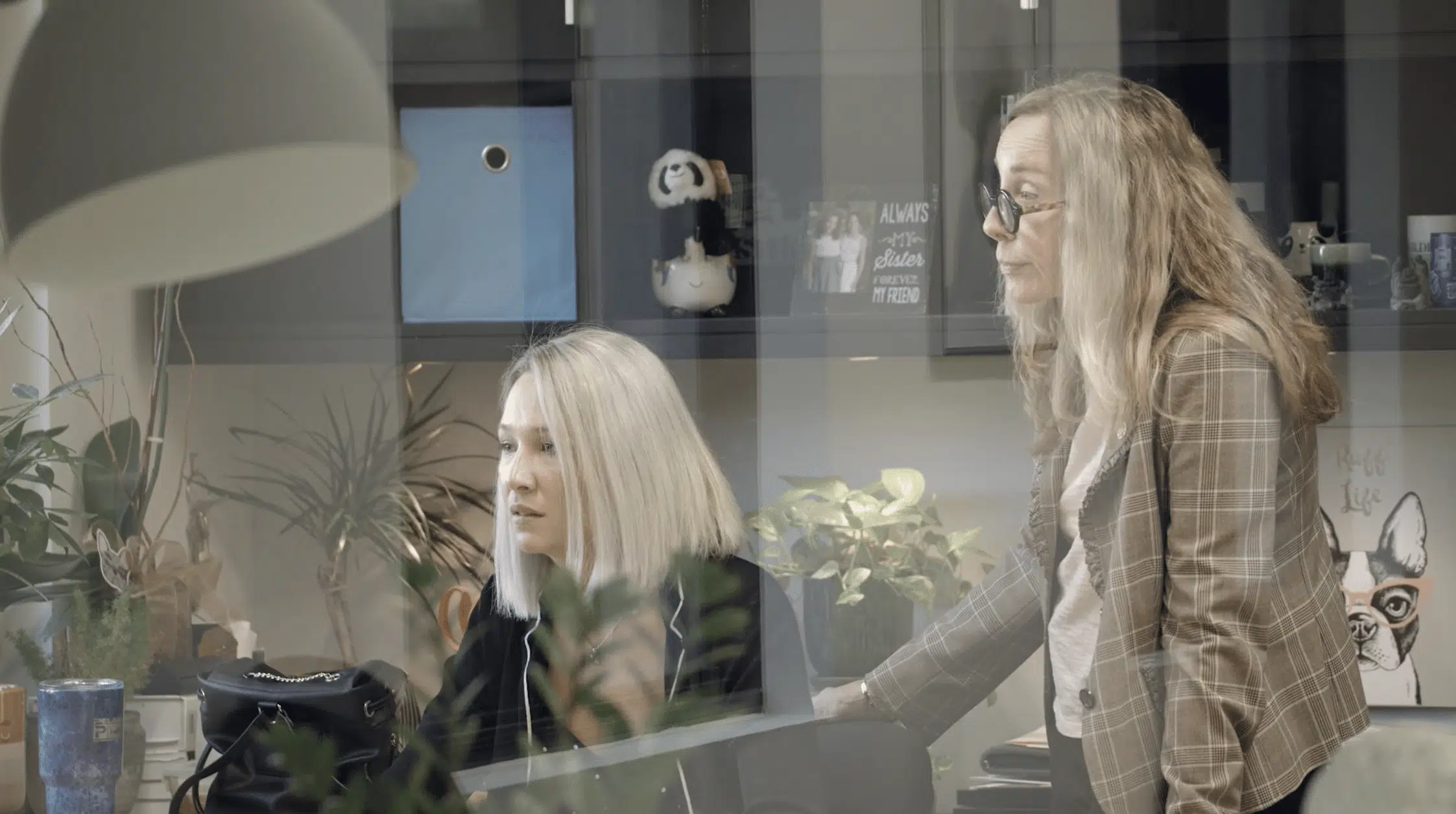 Two women work together in a modern office, seen through a glass wall with reflections. One woman sits at a desk looking at a computer while the other stands beside her, leaning in; shelves, plants, and personal items fill the background.