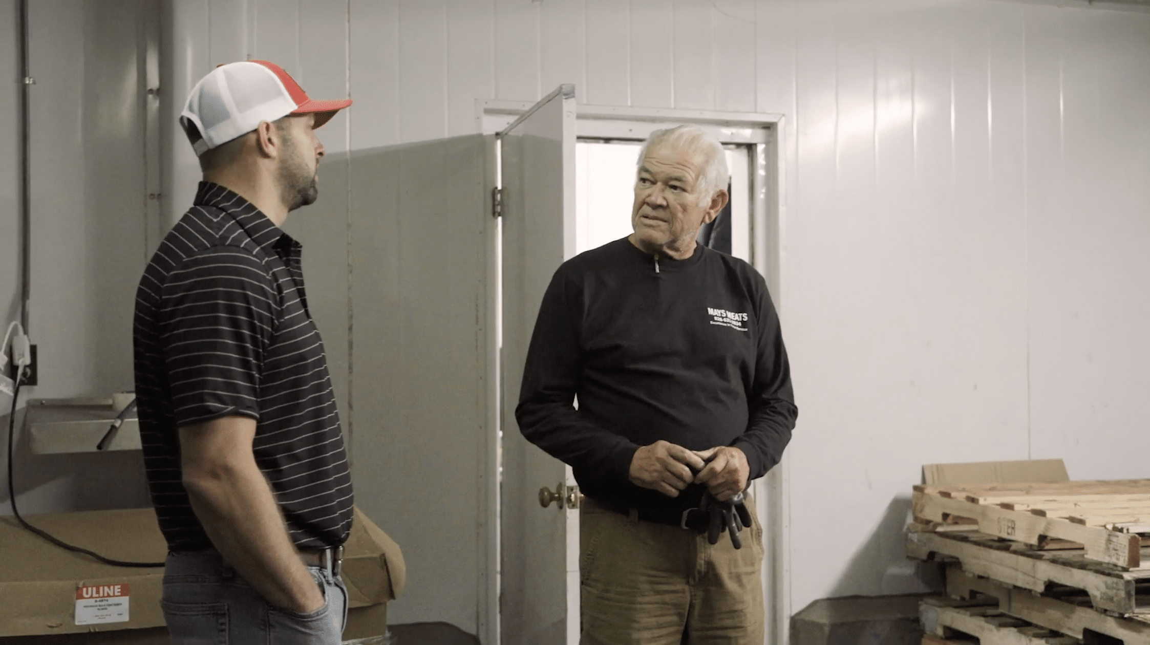 Two men talking, one is wearing a hat and black striped polo shirt, the other is listening and wearing a black long-sleeved shirt and holding a pair of gloves