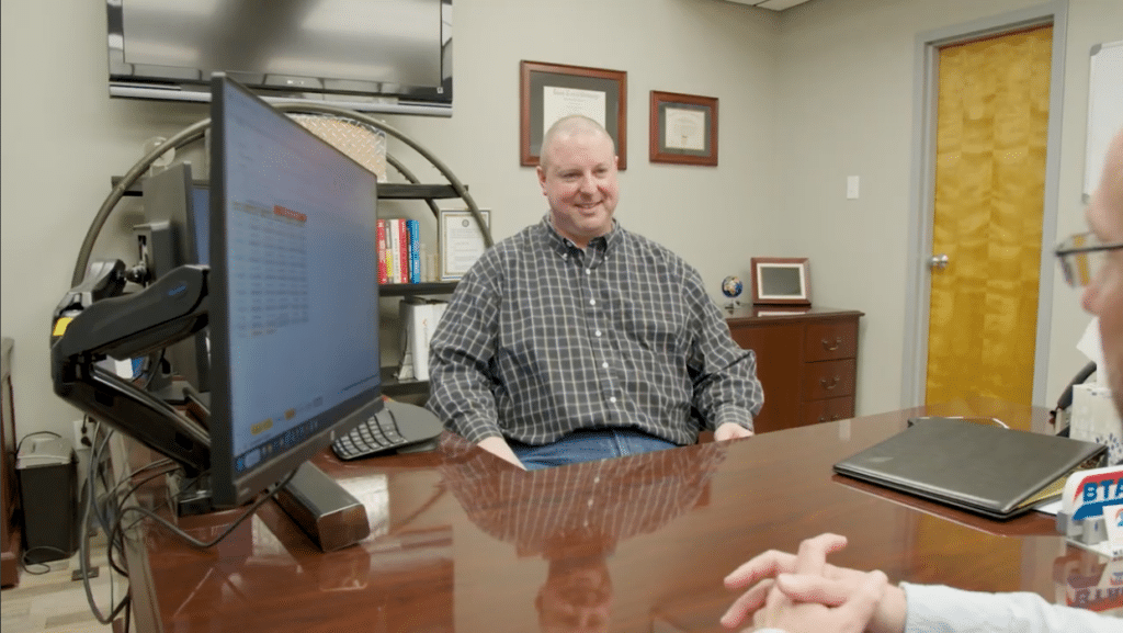 Two men meeting at a desk with a computer screen between them.