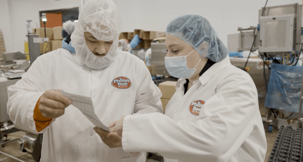 Two employees in protective gear going over a report on the floor of a food manufacturing facility