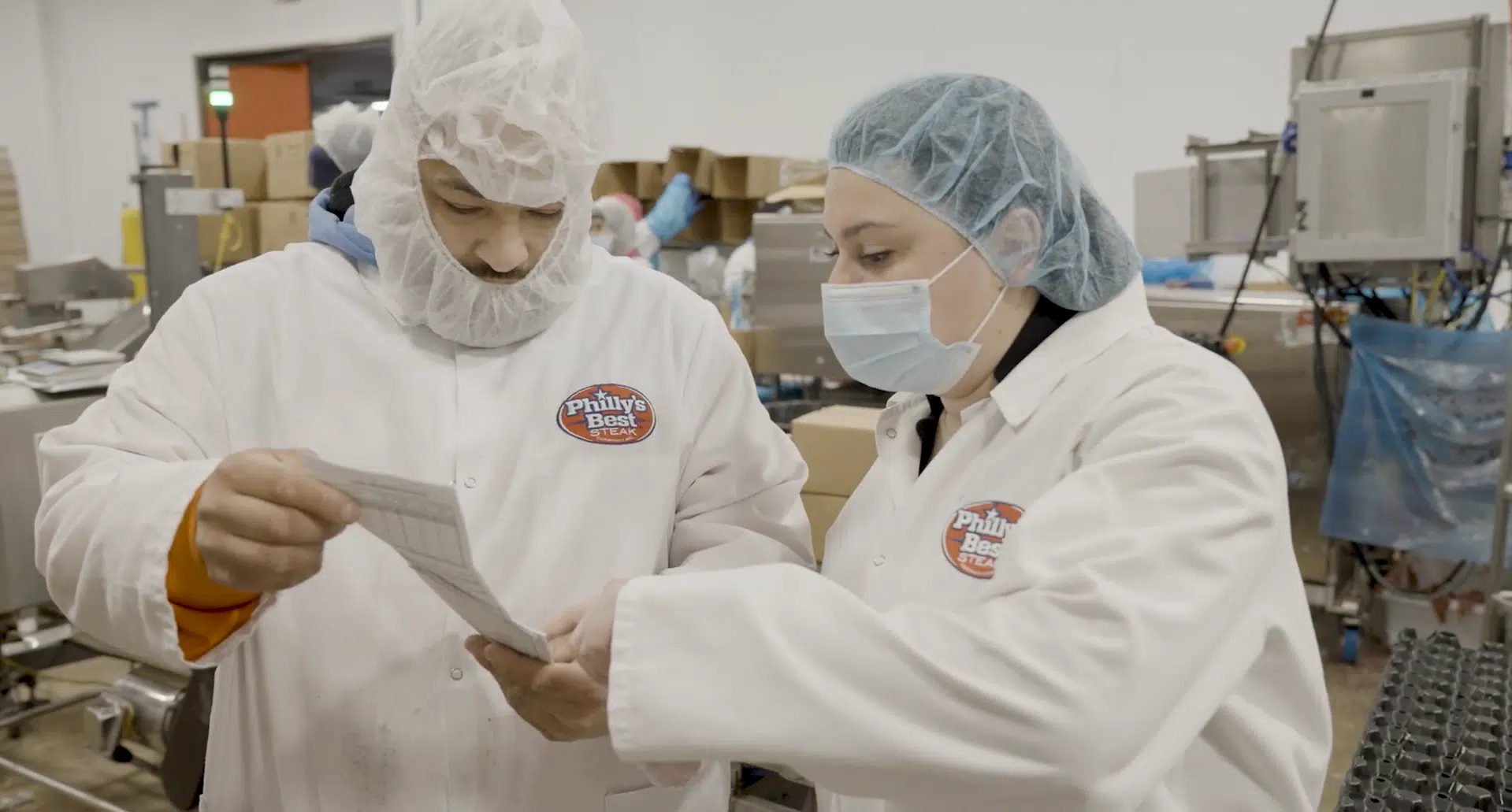 Two employees in protective gear going over a report on the floor of a food manufacturing facility