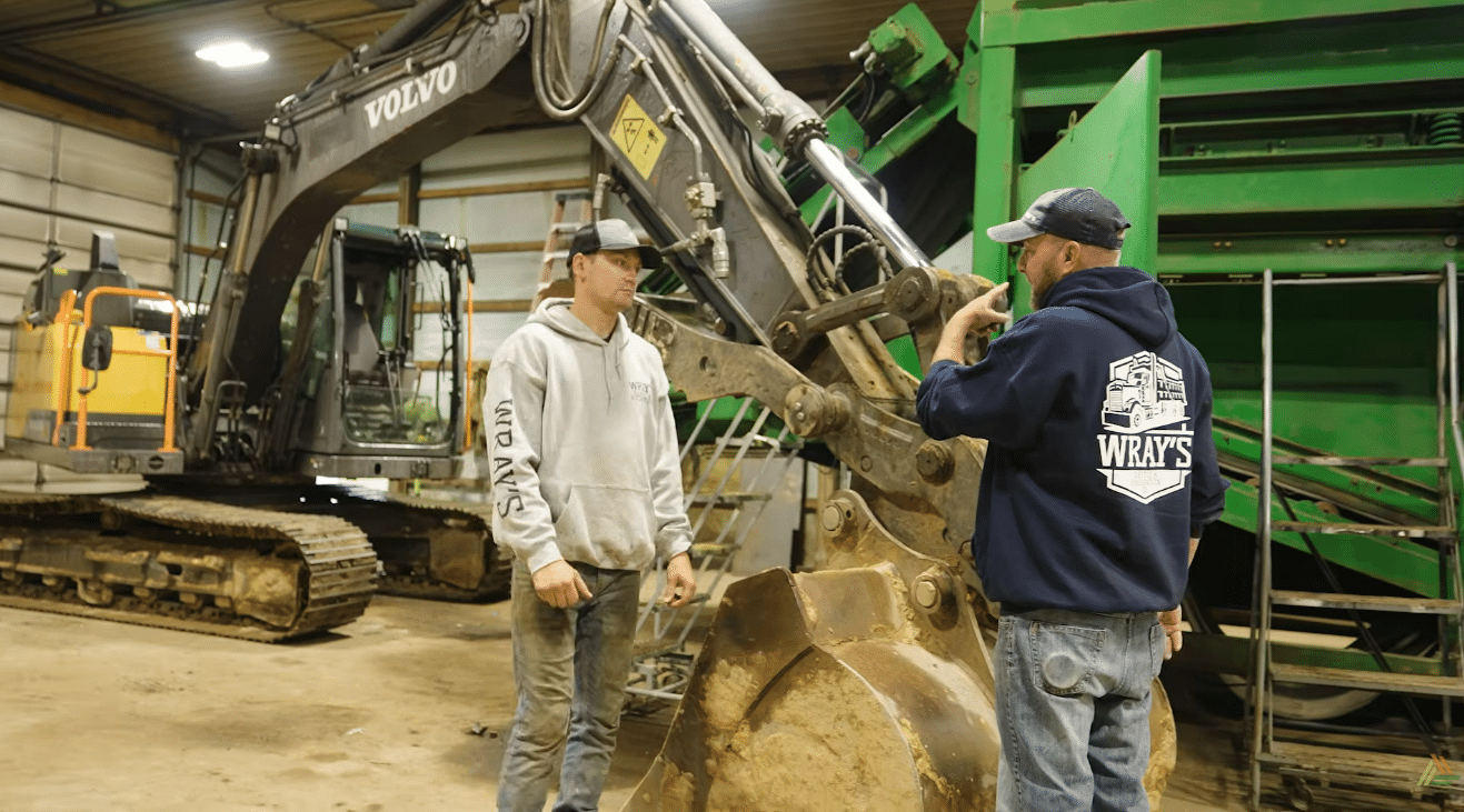 Two workers standing in front of an excavator. One man is giving directions to the other.