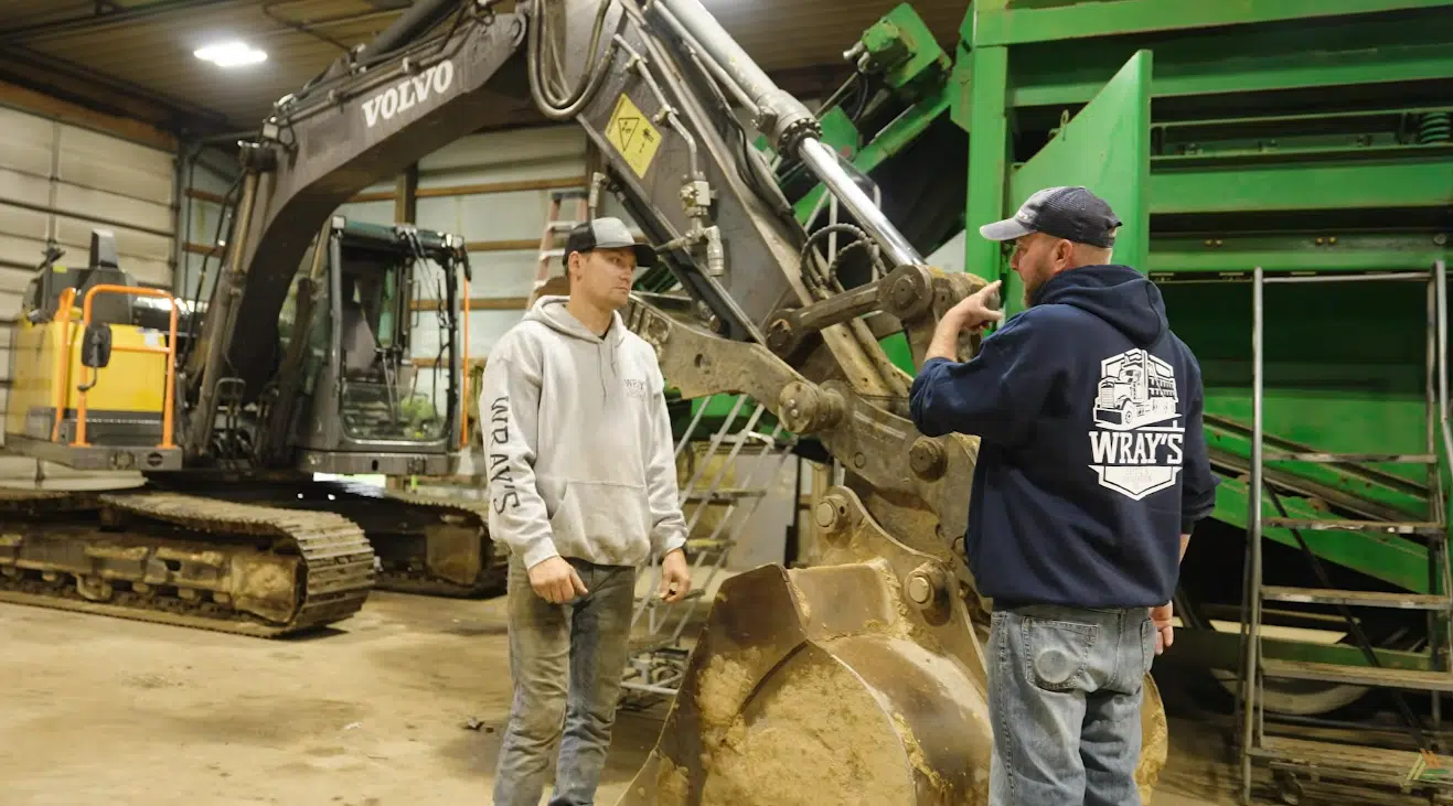 Two workers standing in front of an excavator. One man is giving directions to the other.
