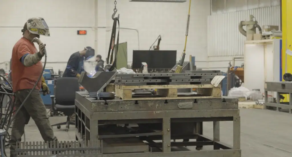 A welder wearing safety gear in a fabrication shop. He is holding tools and working on a large piece of metal.