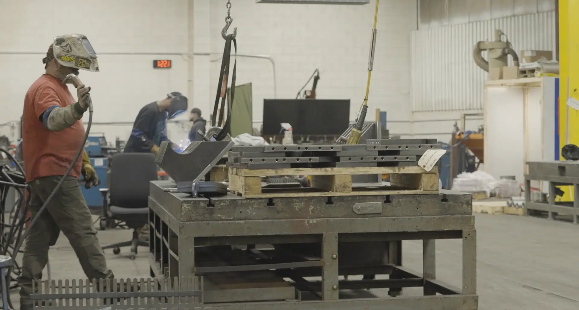 A welder wearing safety gear in a fabrication shop. He is holding tools and working on a large piece of metal.