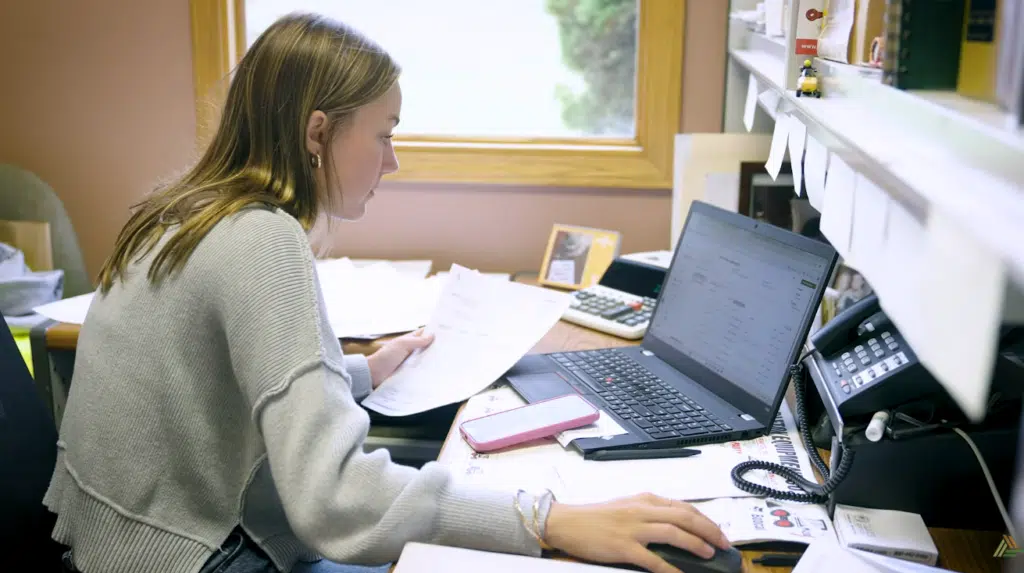 An office employee sitting at a messy desk reviewing her laptop screen.